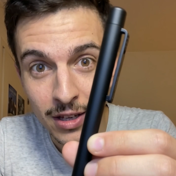 A young man with brown eyes and a mustache holds up a sleek black pen with a clip towards the camera, wearing a light gray shirt against a neutral background.